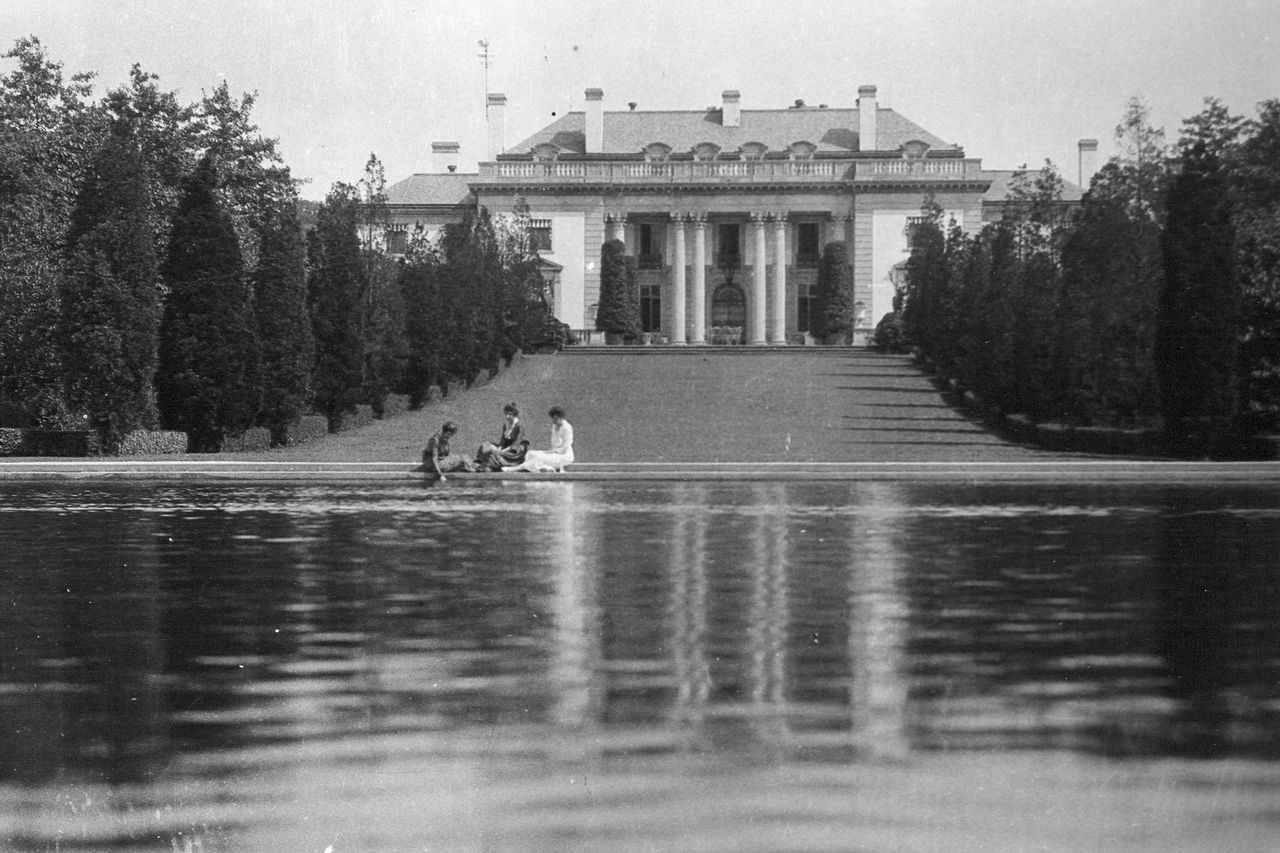 A historical black-and-white photo of three women in the distance sitting by the Reflecting Pool at Nemours Estate with the mansion building in the background. 
