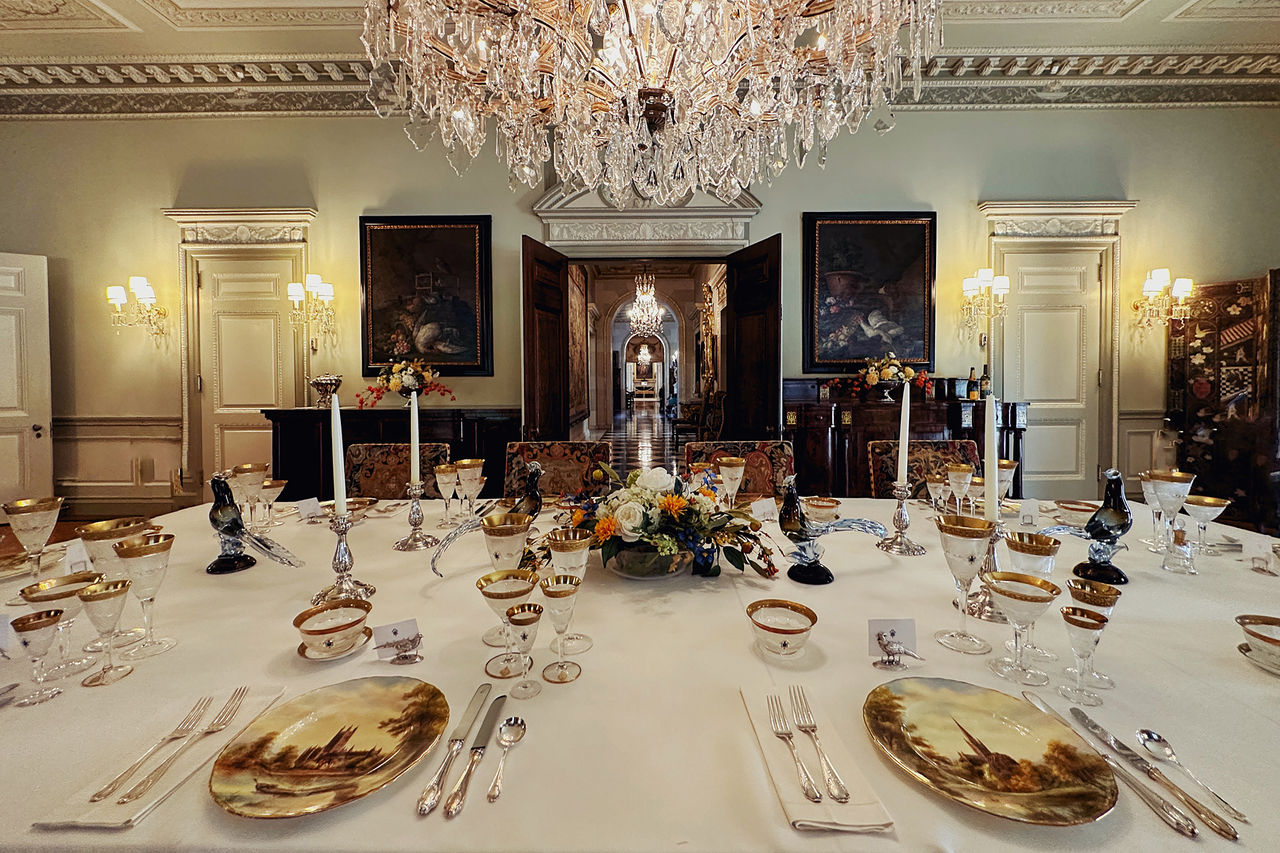 A formal dining table with gold china and place settings, a crystal chandelier, candlesticks on the table and crystal glasses for each guest. 